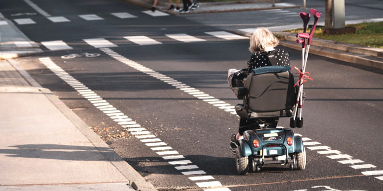 Woman wearing a black and white spotted shirt driving on a street in a motorised wheelchair, with her crutches tied to her wheelchair with a red bow.