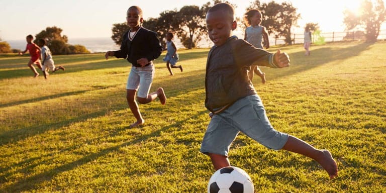A group of young children running, having fun and kicking a soccer ball in a green field with the sun shining in the background.