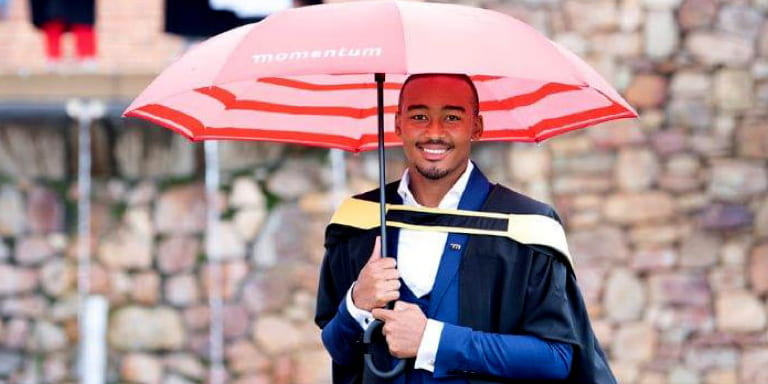 Bathandwa Issac, first Momentum2Exellence bursary graduate, wearing his graduation gown and holding a red Momentum branded umbrella.
