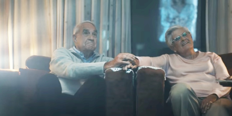An elderly couple is sitting together, holding hands, watching television together.