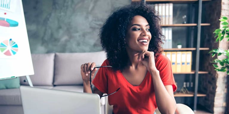 Happy, smiling woman employee wearing a red dress and holding her glasses, seated at her desk in a modern office.