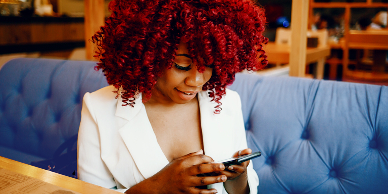 A lady with red hair wearing a white jacket smiling while scrolling on a cellphone.