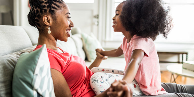 A mum sitting on the bed and her young daughter sitting on top of her belly, looking at each other smiling and holding hands.