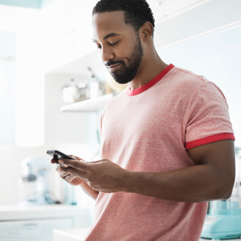 A young muscular man in a red t-shirt standing against a counter in his kitchen reads with a smile on his face about the Momentum Metropolitan Holdings Limited acquisition of Alexander Forbes Insurance.