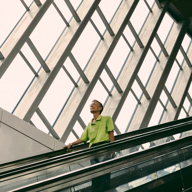 A man in glasses and a green golf t-shirt holds the rails of the escalator with his right hand, as it is coming down.