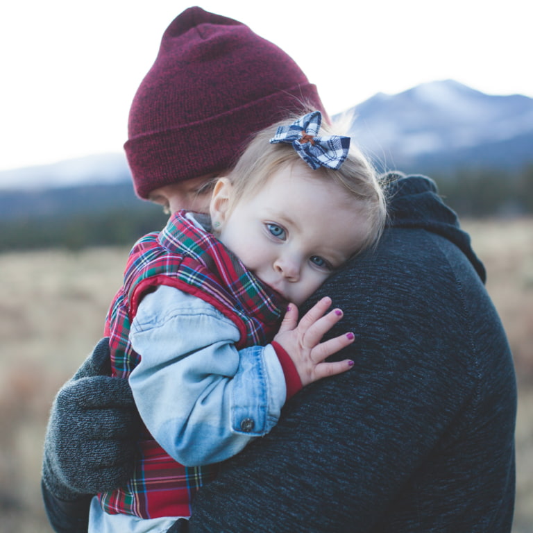 Dad holding his toddler daughter. They're both warmly dressed and wearing beanie hats. Behind them is a mountainous scenery.