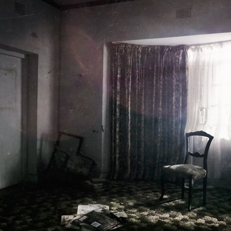 Two chairs in a living room of an abandoned home. One chair has a broken leg with a pile of old newspapers stacked on the floor next to it.