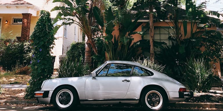 A classic silver grey Porsche parked outside a house with lots of trees.