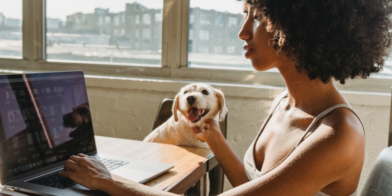 A lady sitting at a disk with one hand on her laptop and the other touching her dog. 