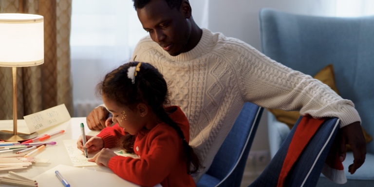 A father sitting with her daughter at a disk as she writes on a piece of paper.