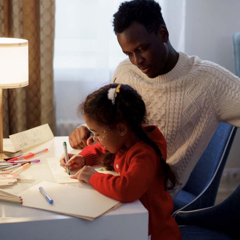 A father sitting with her daughter at a desk as she writes on a piece of paper.