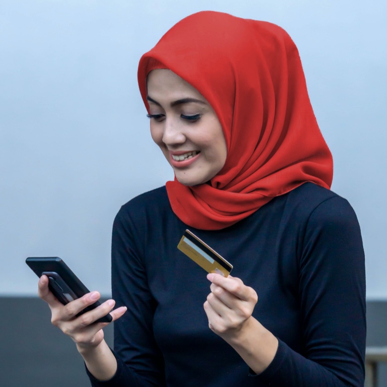 Young lady smiling while holding her cellphone on one hand and a credit card on the other. Wearing a red hajab scarf and navy top. 