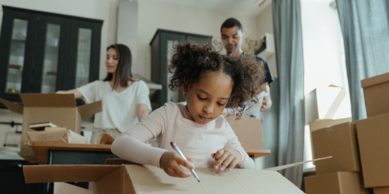 A couple packing boxes to move to their new home while their young daughter marks one of the boxes.