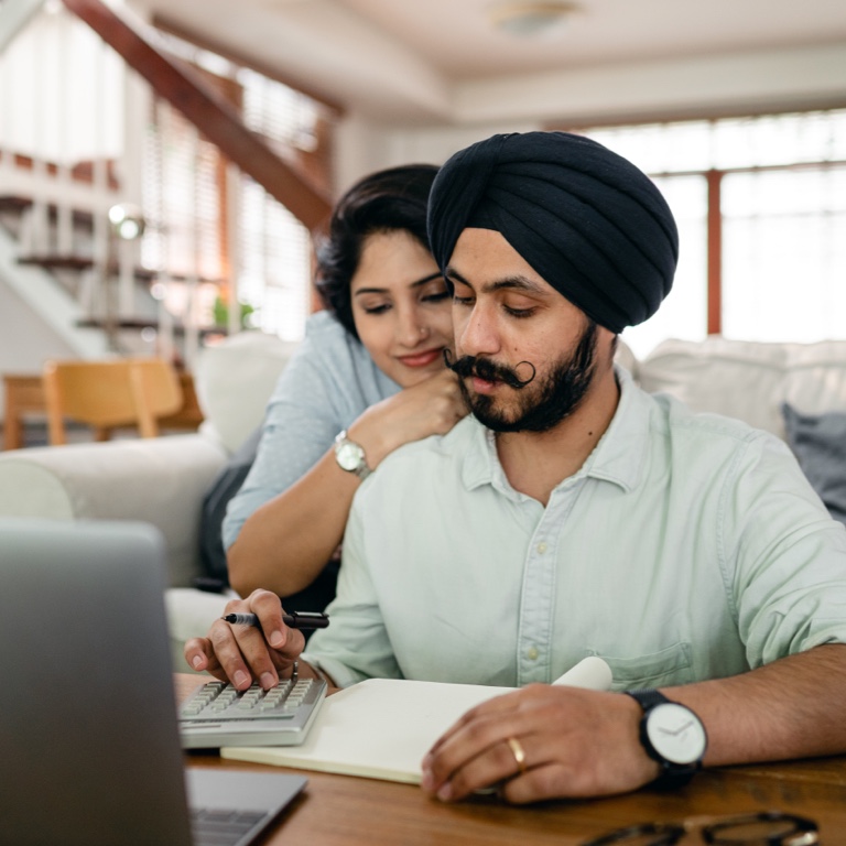 A woman looking over man’s shoulder while he works with a calculator, a pen and paper. There is a laptop in front of them.