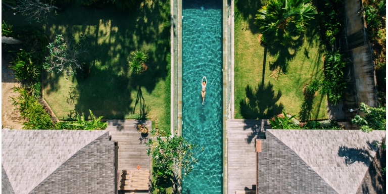 A hovering shot of a women swimming in a pool surrounded by grass, lush trees and grey-tiled roof. 