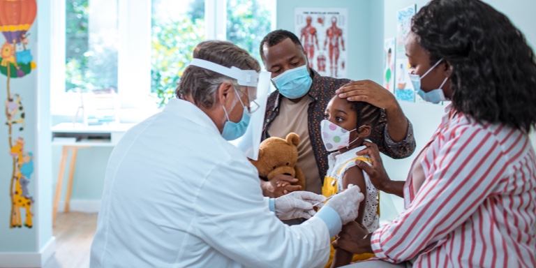 A mother and a father with their daughter in a doctor’s room. The doctor is injecting the little girl and her parents are comforting her.