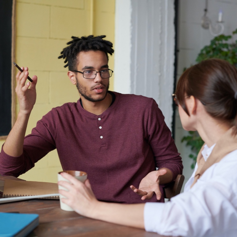 A young man with dreadlocks and a woman sitting at a desk, discussing investments with a laptop in front of them. 