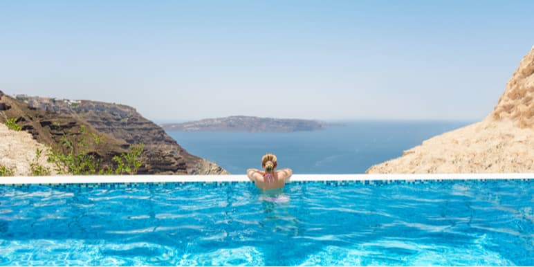 A woman leaning over the edge of an infinity pool overlooking mountains and the ocean. 