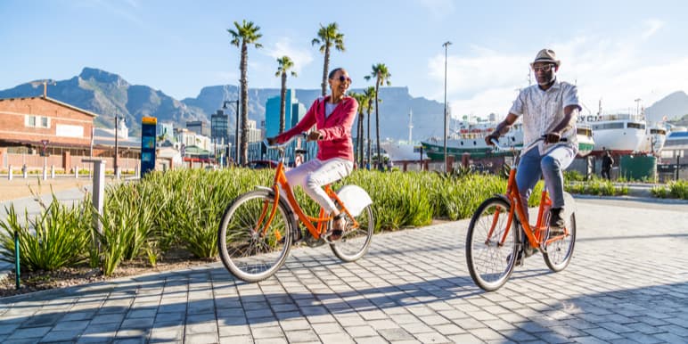A man and a woman riding orange bicycles with a background of The Table Mountain and the harbor. 