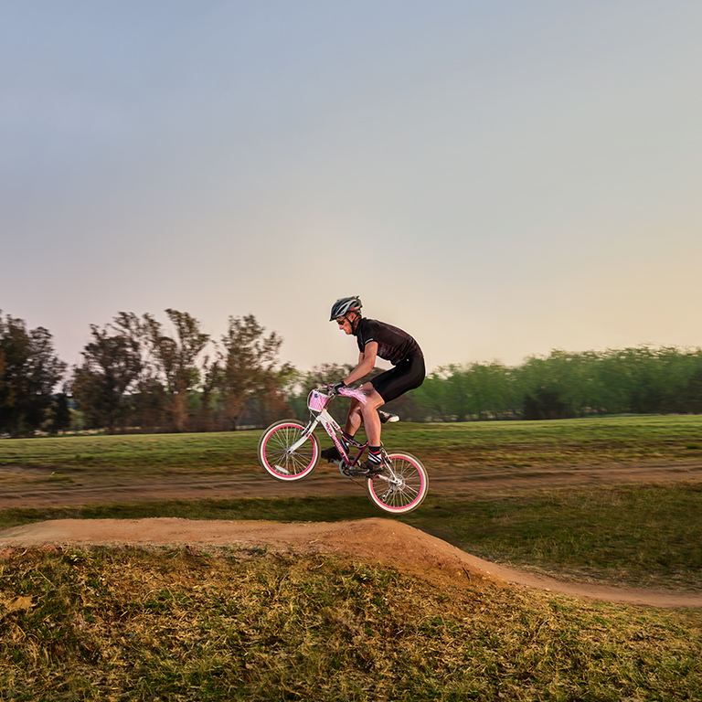 A man in cycling gear participating in a cycling competition with a pink bicycle that is far too small for him. He is pictured going over a sand mound.