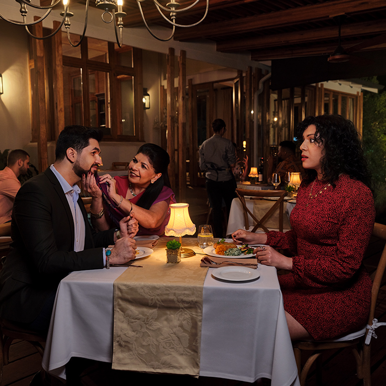 A couple on a date in a restaurant – the female looks at her date confused as his mother is also on the date and wipes crumbs off of her son’s face.
