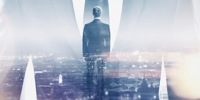 Financial adviser dressed in a smart suit with his back to camera, overlooking a built-up city in the evening