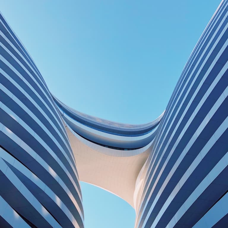 Curved towers of a glass building in a light and dark shade of blue linked together by a sky bridge against the backdrop of a blue sky.