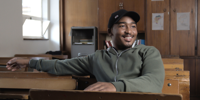 Bathandwa Isaac, a recipient of The M2E Bursary Programme, smiling as he sits at a desk in a school classroom.