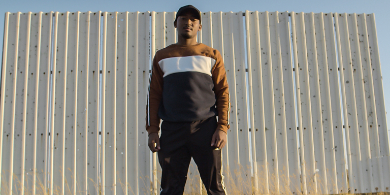 Bathandwa Isaac standing in front of a corrugated iron wall wearing track pants and a striped sweater top.