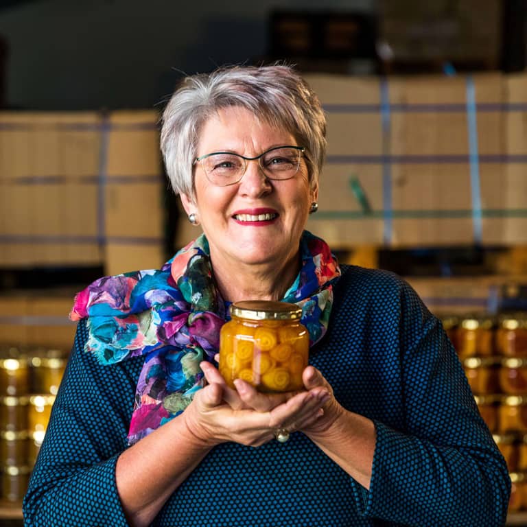 Ina Lessing, entrepreneur and successful business woman standing in front of rows of packed jam jars, proudly holding a bottle of jam.