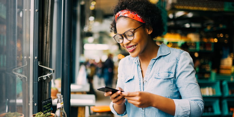 Young lady dressed in a blue denim top, wearing glasses and a red scarf tied around her head smiles as she looks at her cell phone.