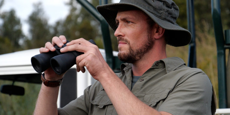 A white man wearing a hat is holding a pair of binoculars.