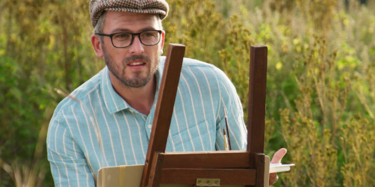 A white man wearing glasses painting in the bush.
