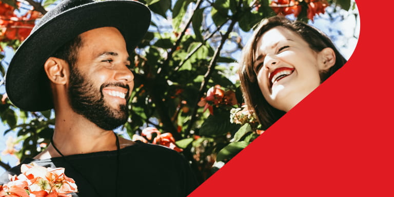 A man smiles while looking at a woman and the woman is laughing with her eyes closed. They’re surrounded by red flowers. 