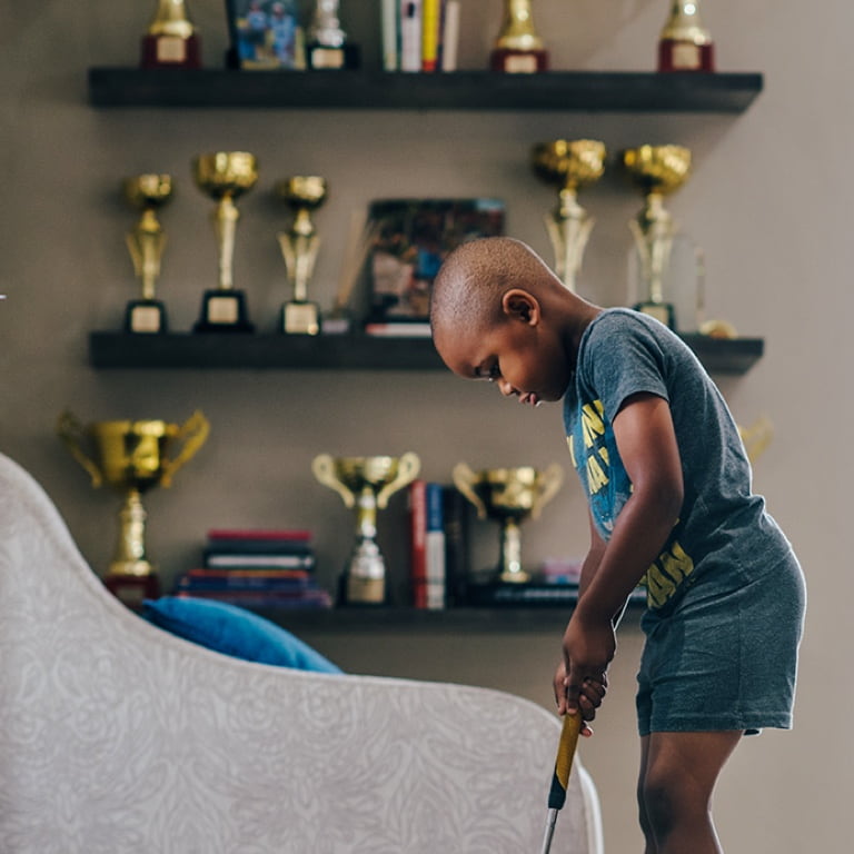 Ngobani Dlamini practicing his putting skills on a practice home kit in front of shelves displaying his trophies.