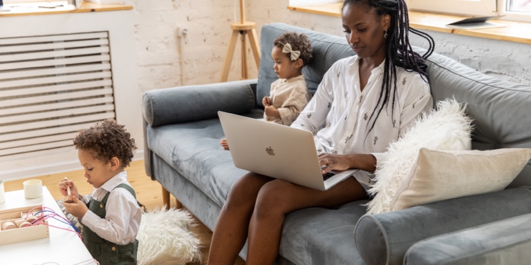 Professional woman sits on her couch working from her laptop while surrounded by her children.