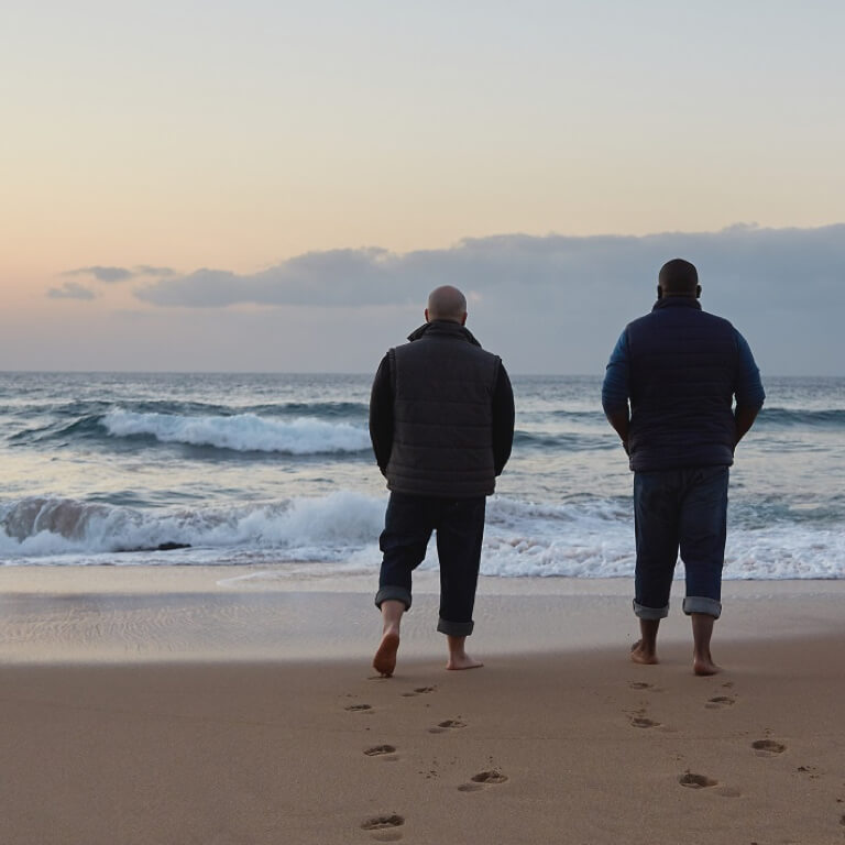 A financial advisor and his client with their backs to camera walking on the beach towards the sea in the early morning.