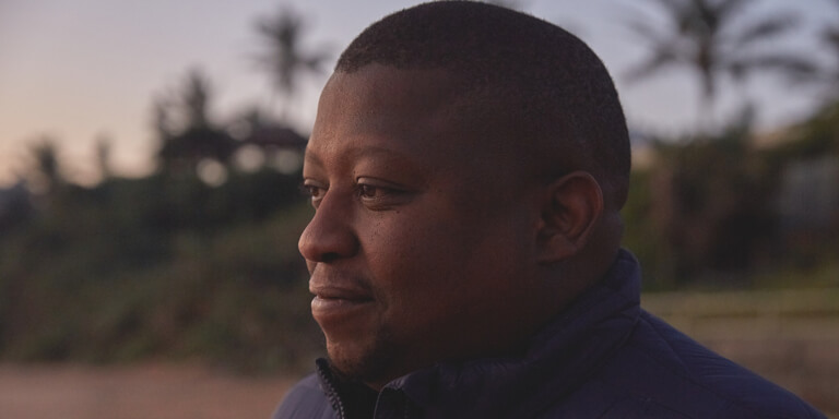 Close up of a mans face who is looking content as he stands on the beach with palm trees in the distance behind him.