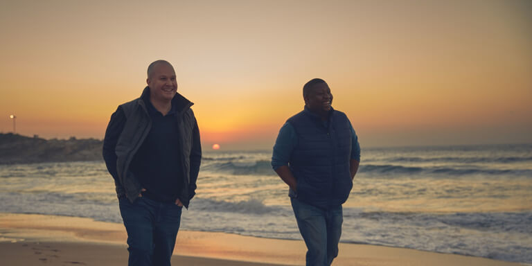 A financial advisor and his client happily chatting and walking along the beach shoreline while the sun sets in the distance.