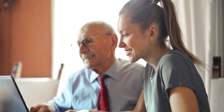 A lady and an elderly man sitting in front of a laptop and smiling with bright daylight coming from the window next to  them. 