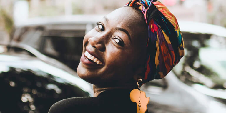 Young black woman turns around to face camera and smile. She is wearing a yellow earing in the shape of Africa and a vibrantly coloured doek on her head.