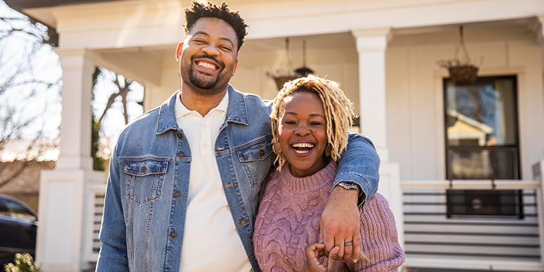 A young couple standing in front of their home’s porch front happy about receiving 15% of their paid premiums back in cash for remaining claim-free for 4 consecutive years with Momentum Rewarder.