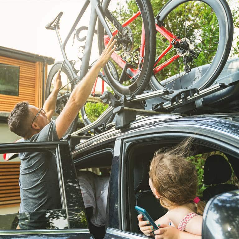 A little girl looking through the window of the passenger seat as her dad secures their bicycles to the roof rack. 