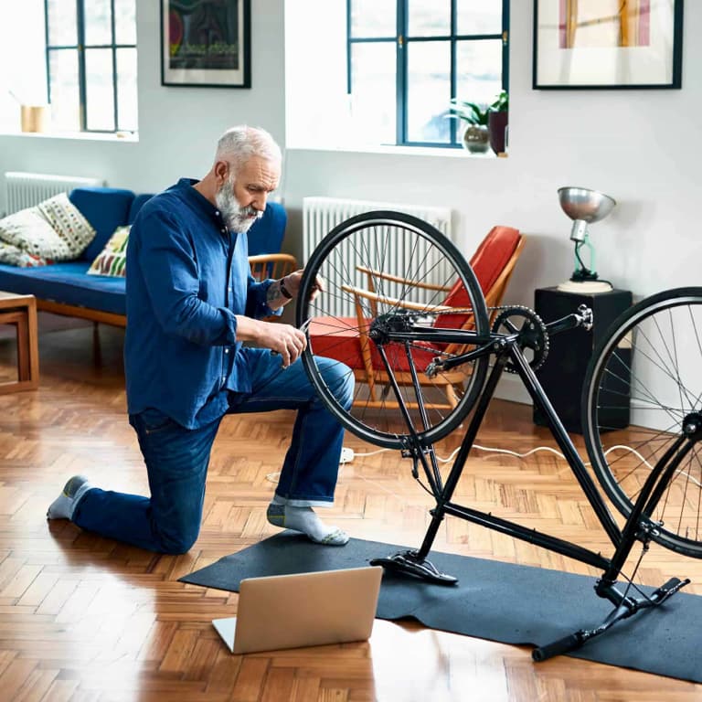 Old man in his blue overalls fixing his bicycle that's placed upside down in his living room.
