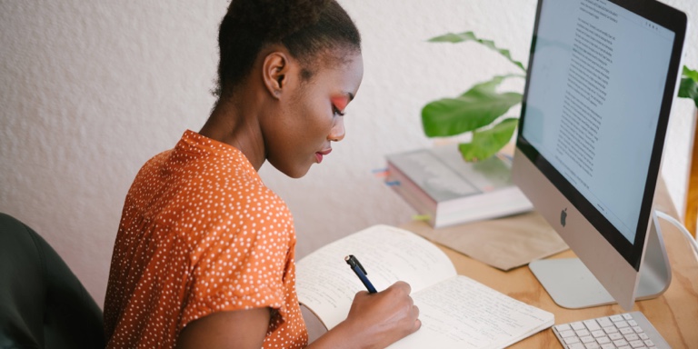 A business owner making notes in her notebook while in front of her desktop computer considering solutions to make sure her business can continue to make money if she were to become critically ill, disabled, or die.