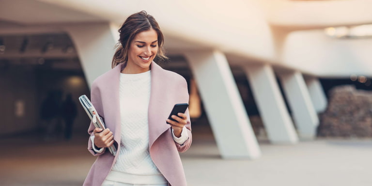 Lady wearing white jeans, white jumper, and a pink jacket walks out of a building holding her folder in one hand while looking at her cell phone reading up about Business Continuity Assurance products for her business.
