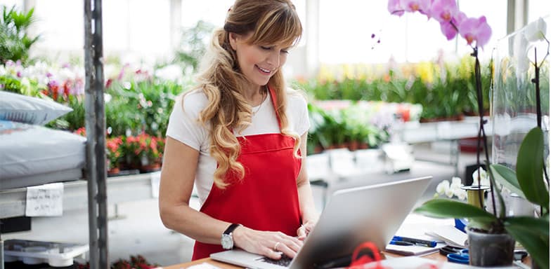 A lady standing in a nursery with flowers in the background working on a laptop, wearing a red apron. 