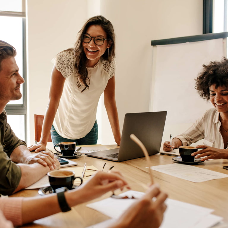 Four colleagues with black coffee mugs laughing around a table at a work meeting.