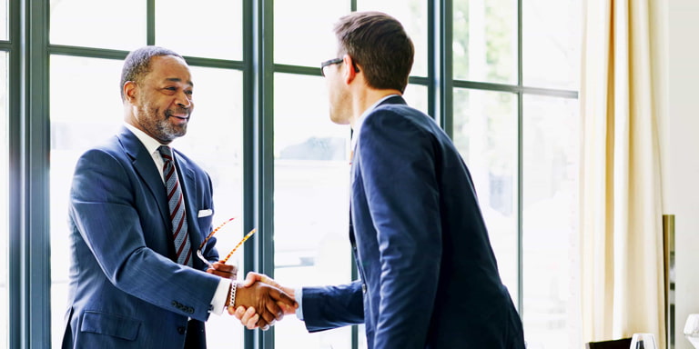 An older male financial adviser shaking hands with a male business executive in a boardroom.