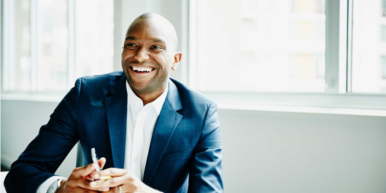 A laughing young African male professional at a desk,  in a suit with a mobile phone.